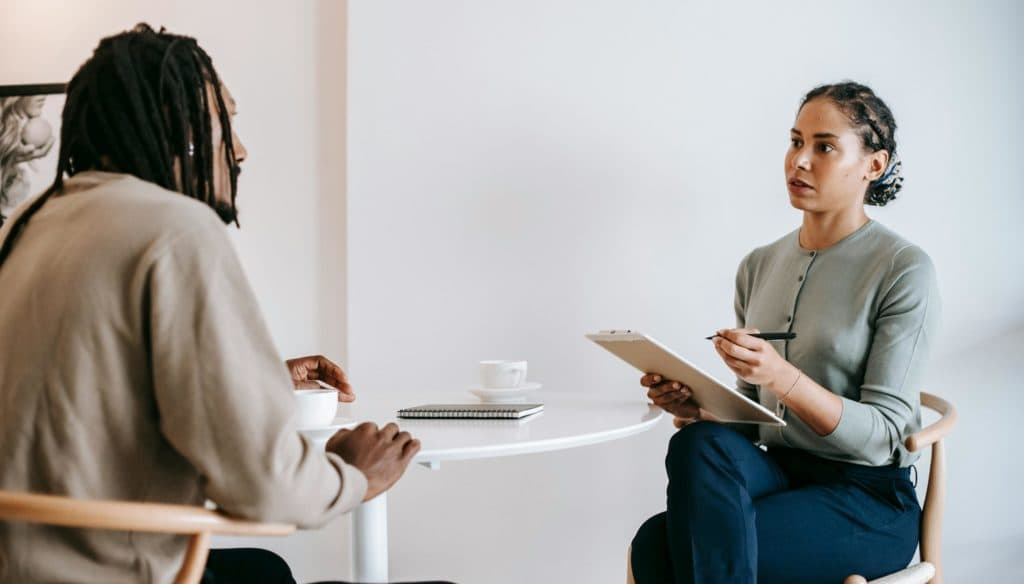 Two people seated at a table. One person has a clipboard. Its a professional setting indicating an employee coaching session as part of an employee development coaching cadence.